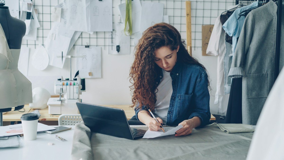 A designer sketching at her workshop table beside a laptop